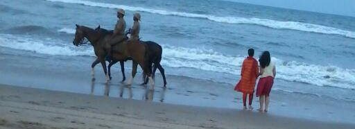 Two horseback riders in uniforms trotting along a beach while two people hold hands and walk near the water's edge.